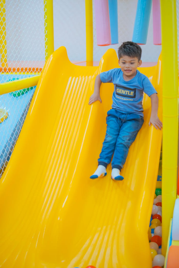 About Boy sliding down a yellow slide in an indoor playground with colorful balls below.