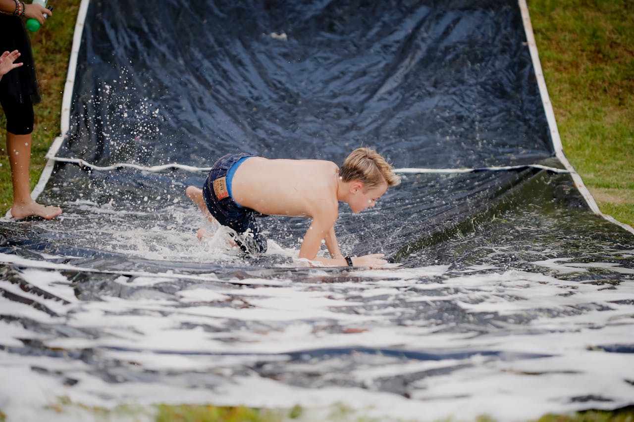 Services A young boy having fun on a slip and slide outside during a sunny day, splashing through water.